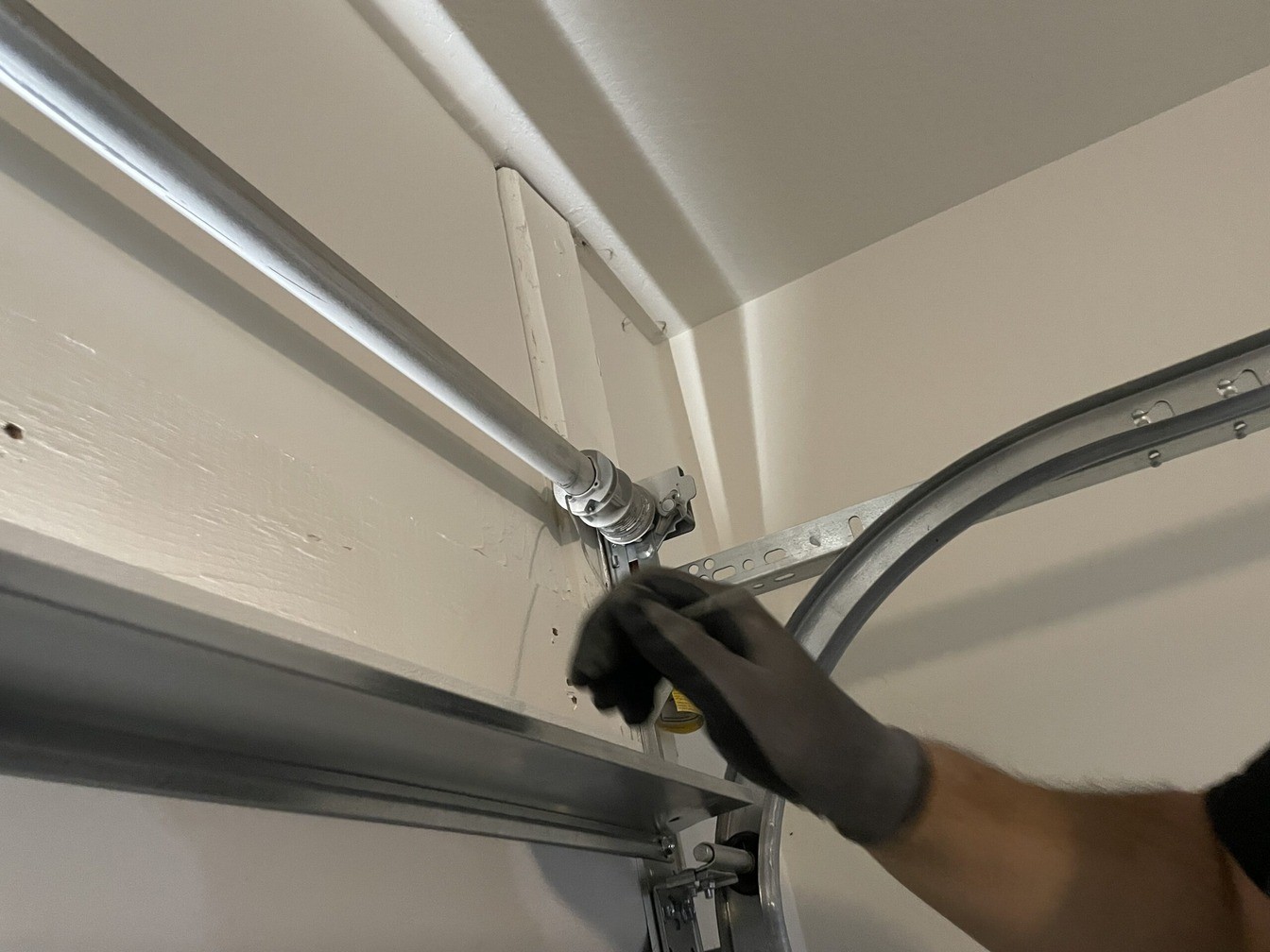 A gloved hand works on the cable hardware where the garage door track curves overhead.